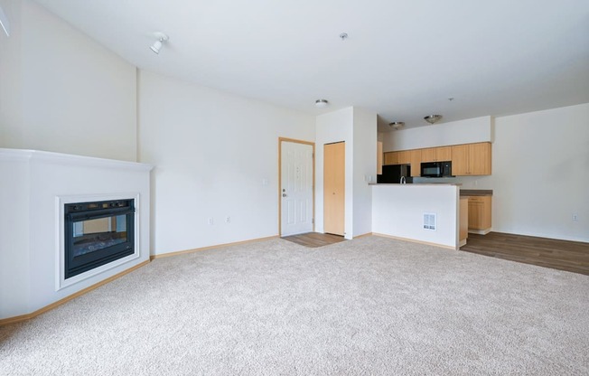 Spacious Soho living room with a fireplace, carpeting, and a kitchen in the background at Abbey Rowe Apartments in Olympia, WA