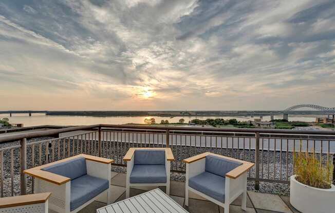 Two chairs and a table on a balcony overlooking a river at sunset.