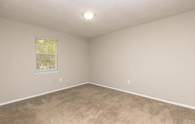 A vacant room with beige walls and a light-colored carpet. There is a single window with blinds allowing natural light to enter. The ceiling is plain with a central light fixture. The room appears spacious and simple, suitable for various uses.