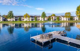 A serene lake with a dock and chairs in the foreground and apartment buildings in the background.