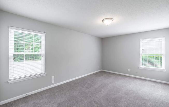 A room with a carpeted floor and two windows with blinds.