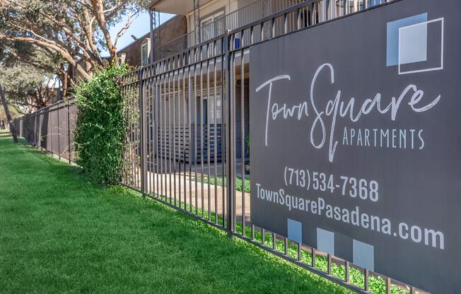 Sign for Town Square Apartments featuring the name, contact number (713-534-7368), and website (TownSquarePasadena.com). The sign is positioned on a black metal fence with greenery and a well-maintained grassy area visible in the background.