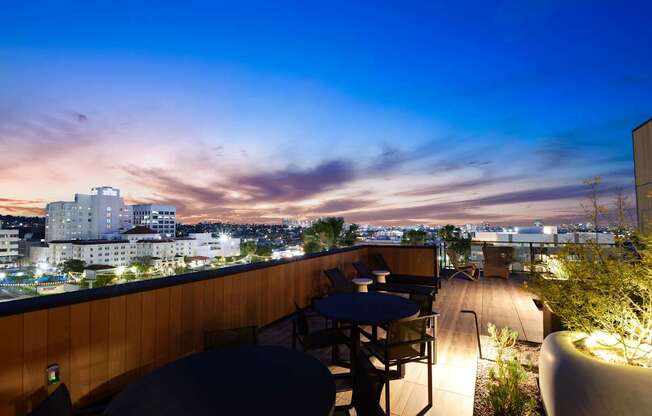 A rooftop patio with tables and chairs overlooking a cityscape at dusk.