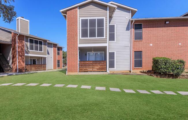 A view of an apartment complex featuring two buildings. One building has a modern grey and brick exterior with a balcony, while the other is predominantly brick. Well-kept green lawns and a stone pathway connect the two buildings under a clear blue sky.