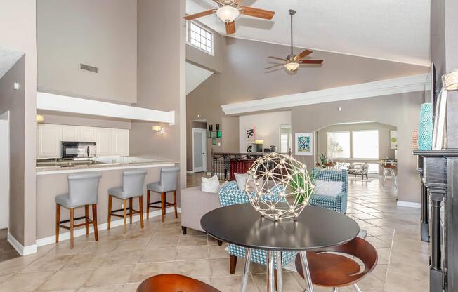 A spacious and modern living area featuring a round dining table with brown chairs, a light-colored sofa, and decorative elements. The kitchen area is visible with bar seating. Large windows allow natural light, and the room has a neutral color palette with ceiling fans and tiled flooring.