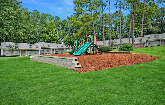 A playground with a green slide in the middle of a grassy area.