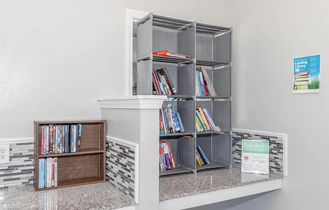 Bookshelf displaying a variety of books and DVDs in a neatly organized area. The shelf has multiple levels filled with colorful book spines, while a smaller shelf to the left holds DVDs. A sign indicates a "Free Books" lending library. The backdrop features a stylish mosaic tile design.