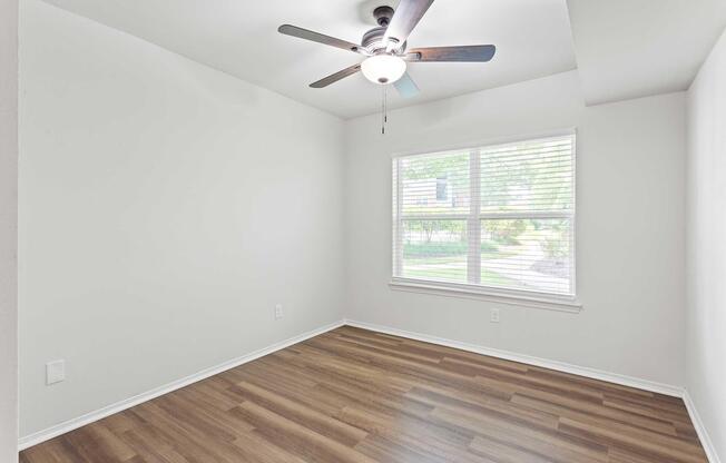 Empty room with light-colored walls and a ceiling fan. There is a window with blinds allowing natural light to enter, revealing a green outdoor view. The flooring is a wood laminate, and the space appears clean and well-maintained, suitable for various uses, like a bedroom or office.