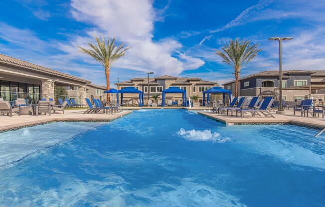 A bright outdoor swimming pool surrounded by lounge chairs and cabanas, set against a backdrop of palm trees and modern buildings. The sky is clear with scattered clouds, creating a vibrant and inviting atmosphere. Ideal for relaxation and leisure.