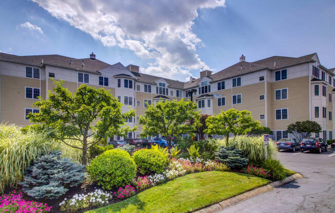 View of community with lush and colorful landscaping with apartment building parking lot of cars