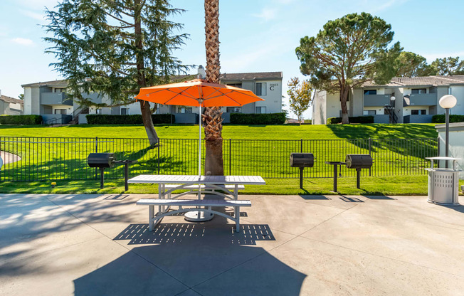 A picnic table with an orange umbrella is in the middle of a concrete area with a tree and mailboxes in the background.
