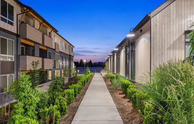 A long walkway separates apartment buildings with green shrubbery on either side.