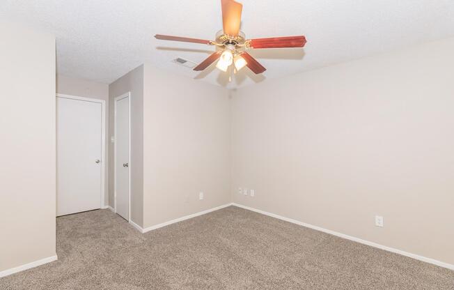 A spacious, empty room with light beige walls and a ceiling fan with wooden blade accents. The floor is covered in light brown carpet. There is a closed door on the left and a closet door on the right, creating a clean and minimalist look, ideal for personalization.