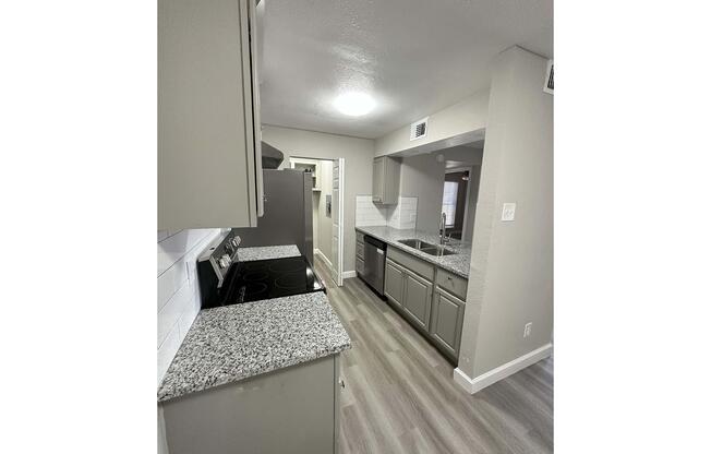 A modern kitchen featuring gray cabinets, a granite countertop, and stainless steel appliances. The space includes a stove, refrigerator, and a sink with a view of an open floor plan. The walls are painted in a light color, and the flooring is a light wood laminate, giving a bright and spacious atmosphere.