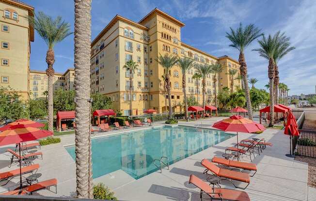 A pool area with red umbrellas and chairs in front of a tan building.