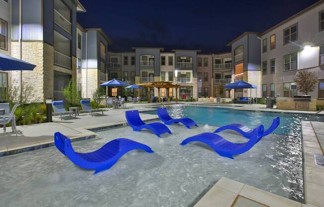 an outdoor pool with blue chairs and an apartment building at night