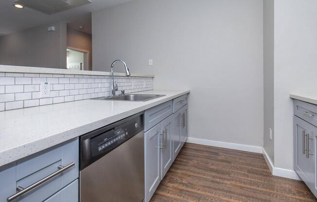 A modern kitchen featuring a clean countertop with a stainless steel sink and a dishwasher. The cabinets are light blue with sleek metal handles, and there is a white subway tile backsplash. The flooring is a warm wood tone, creating a welcoming atmosphere.