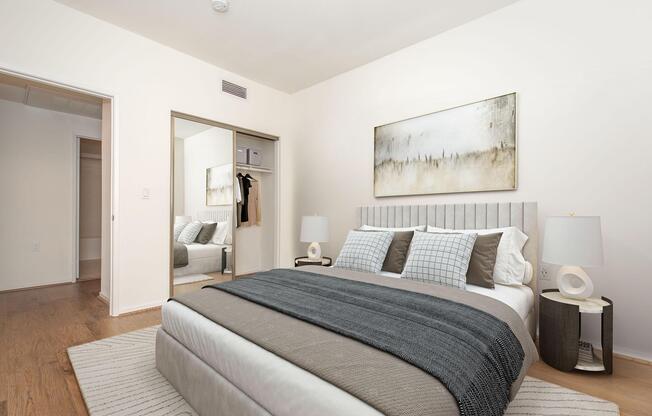 A modern bedroom featuring a queen-sized bed with gray bedding and decorative pillows. Two bedside lamps are on either side of the bed. A large abstract painting hangs above the headboard. There's a mirrored closet reflecting the room and a light-colored rug on hardwood flooring. Soft, neutral wall tones complete the aesthetic.
