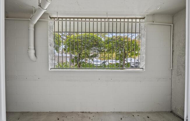 A view from inside a room looking out through a barred window. The window frames greenery and parked cars outside. The walls are bare and painted white, and the floor appears to be concrete. Natural light enters the space, highlighting the window bars.