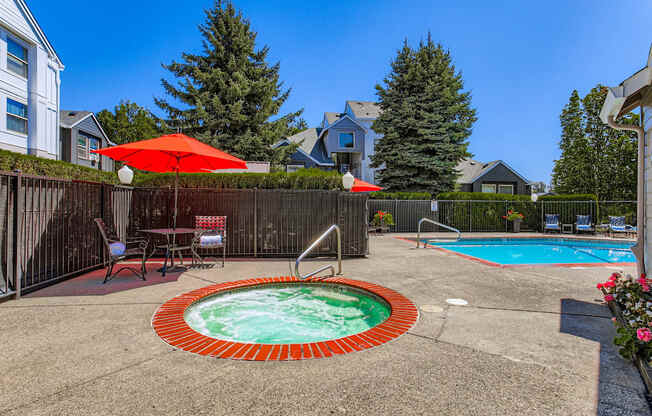 A hot tub sits in the middle of a backyard with a red umbrella and chairs around it.