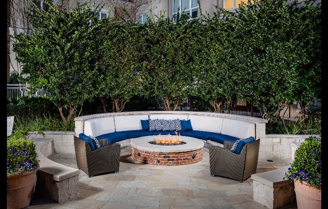 A round banquette couch and two wicker armchairs around a round stone fire pit with trees in the background.
