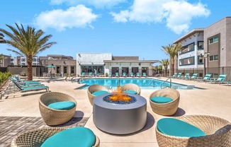 uxurious apartment complex showcasing a stylish outdoor pool area with cozy wicker seating and a modern fire pit, surrounded by palm trees under a clear blue sky.