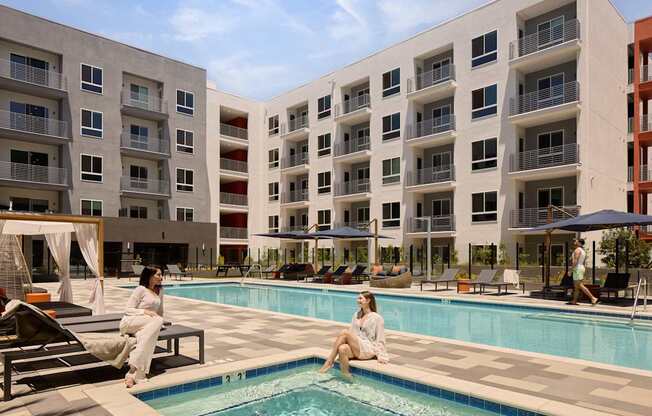 Two women are sitting by a pool in front of apartment buildings.