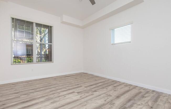 A bright and spacious empty room with light-colored wooden flooring, featuring two windows with blinds. One window is large and extends to the floor, while the other is smaller and higher up on the wall. The walls are painted in a light color, and the ceiling has a modern fan.
