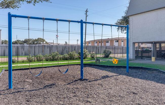 A playground featuring a blue swing set with two swings, surrounded by a black rubberized surface. In the background, there are residential buildings and a fenced area. The setting is sunny, with clear skies and a few trees nearby, suggesting a pleasant outdoor space for children to play.