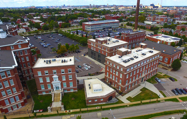 A large brick building with a tall chimney is surrounded by other buildings and a parking lot.