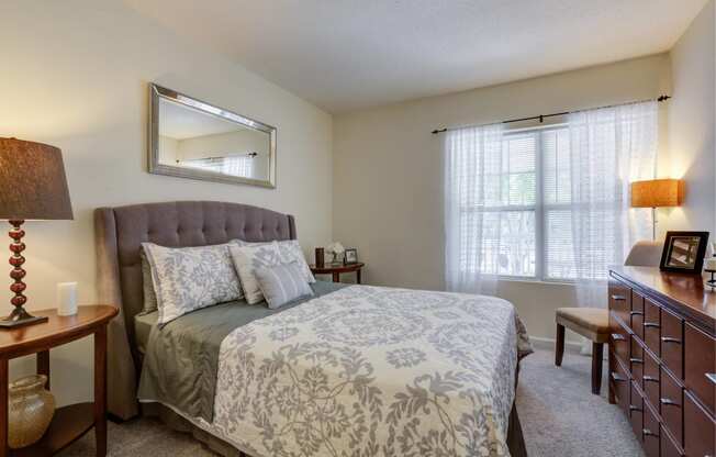 Furnished bedroom with plush carpeting and oversized window with mini-blinds. Room includes queen-sized bed, two nightstands, chair, lamp and horizontal dresser. at Summit Place apartments in Methuen,