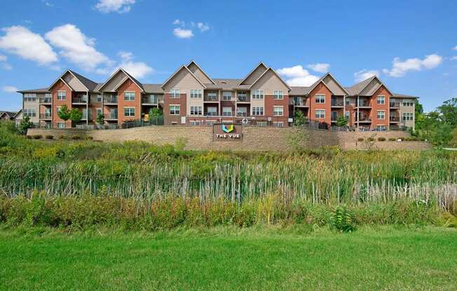 A scenic exterior view here at Vue at Pinnacle Park with multi-story apartment buildings, private balconies, landscaped grounds, and a pond with natural greenery creating a peaceful community setting.