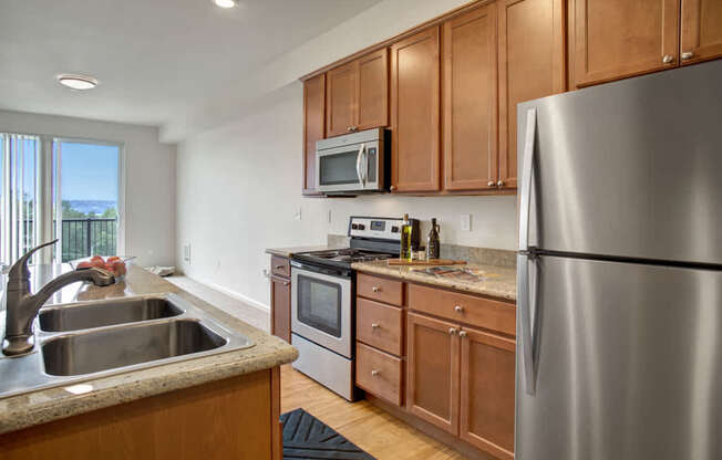 A contemporary kitchen here at Heritage Hills showcasing warm wood cabinetry, granite-style countertops, and stainless steel appliances including a refrigerator, range, and microwave. The space features a double-basin sink with gooseneck faucet, wood-style flooring, and a galley layout that opens toward a dining area and balcony with natural light and scenic views.