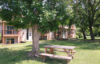 A wooden picnic table is in the foreground of a grassy area with a tree and apartment buildings in the background.