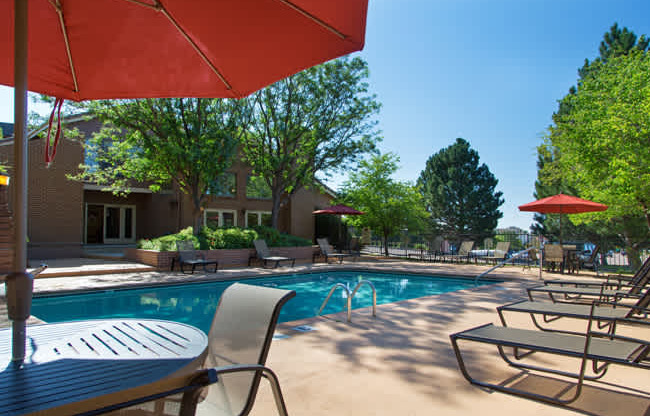 A bright outdoor pool area here at Eagle Ridge featuring a rectangular swimming pool with clear blue water, surrounding concrete deck, multiple lounge chairs arranged along the poolside, patio tables with red umbrellas, mature trees providing shade, and a clubhouse building visible in the background under a sunny blue sky.