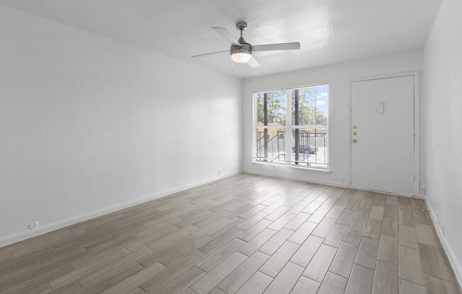 A spacious, bright living area with light-colored walls and tiled flooring. The room features a ceiling fan and a large window allowing natural light to enter. A door leads to the outside, and there is minimal furniture present, creating an open and airy atmosphere.