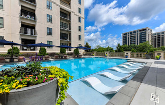 A large swimming pool with a concrete edge and a flower bed in front of a multi-story apartment building.