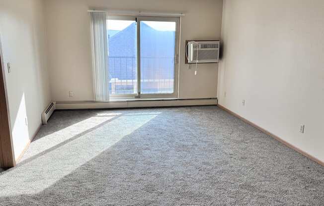 A room with a carpeted floor, a window with blinds, and a wall-mounted air conditioning unit.