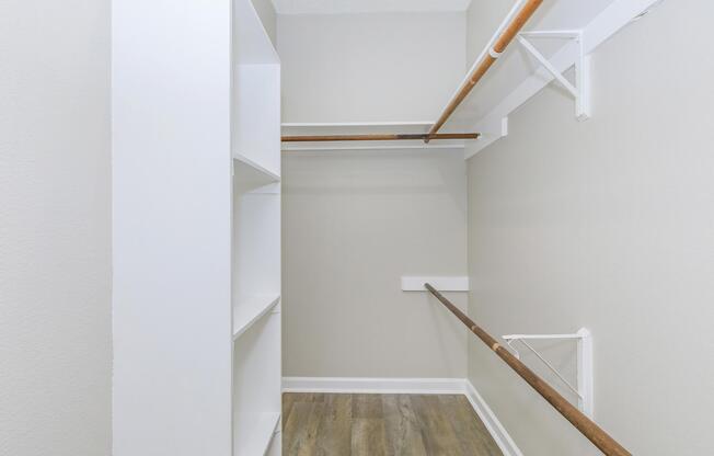 A well-organized closet with white shelves on the left and two wooden hanging rods for clothes on the right. The walls are painted in a light gray color, and the flooring is a wood laminate. The space appears clean and spacious, suitable for storage and organization.