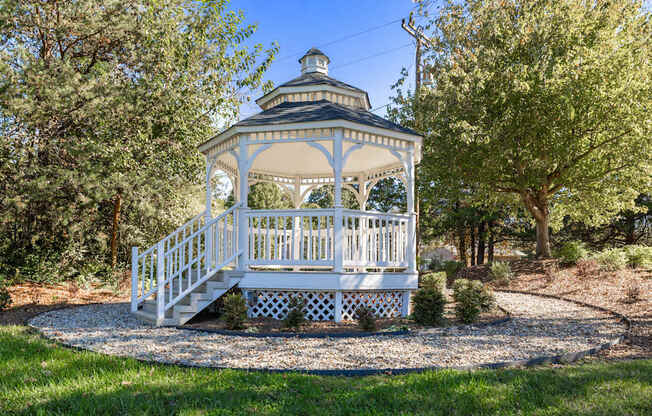 A gazebo with a white railing and steps is surrounded by trees.