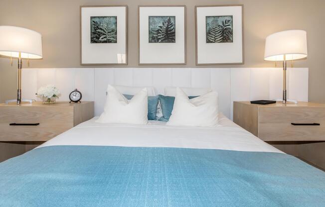 A modern bedroom featuring a neatly made bed with light blue and white bedding. There are two bedside tables with lamps, a clock, and decorative items. Above the bed, three framed prints of ferns are displayed on the wall, creating a serene and stylish atmosphere.