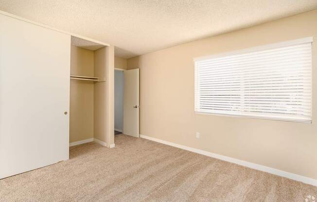 A room with a carpeted floor, a window with blinds, and a door leading to another room.