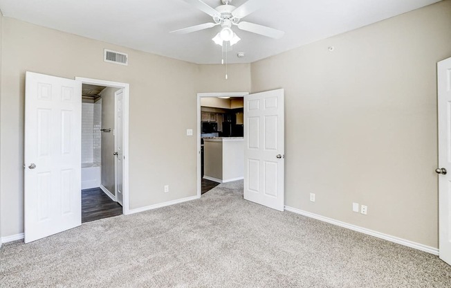 Spacious bedroom at Saxony at Chase Oaks Apartments in Dallas, TX, featuring beige walls, plush carpeting, ceiling fan, and direct access to a bathroom and kitchen.
