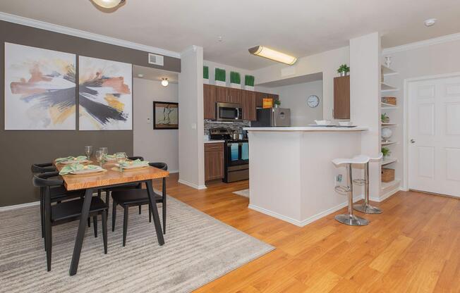 Modern kitchen and dining area showcasing a wooden dining table set for four, with green dishware. The kitchen features dark cabinetry, stainless steel appliances, and a sleek countertop. Decor includes wall art with abstract designs and a rug on hardwood flooring, creating a warm and inviting atmosphere.