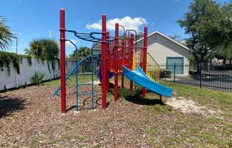 Red and blue playground set in a bed of mulch surrounded by a black fence with buildings and palm tree in the background