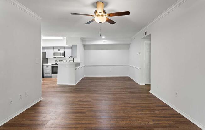A spacious and modern living area featuring a ceiling fan, light-colored walls, and hardwood flooring. On one side, there is an open kitchen area with stainless steel appliances and a breakfast bar. The layout offers a bright and inviting atmosphere, suitable for various furniture arrangements.