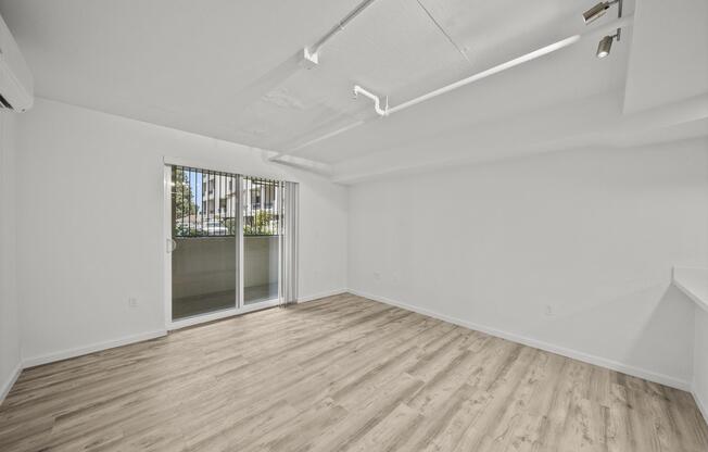 A spacious, empty room with light-colored walls and wooden flooring. There are large windows with vertical blinds, letting in natural light. The room features modern lighting fixtures and a sliding glass door leading to a balcony or outdoor area. The overall atmosphere is bright and inviting.