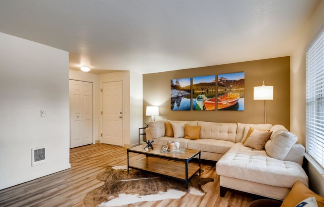 Apartments in Happy Valley, OR, for Rent - Sunnyside Village - Spacious Living Room with Wood-Style Flooring, Animal Skin Rug, Cream-Colored Sectional Couch, Wooden Coffee Table, Floor Lamp, Side Table with Lamp, White Walls with a Beige Accent Wall, and Large Window.