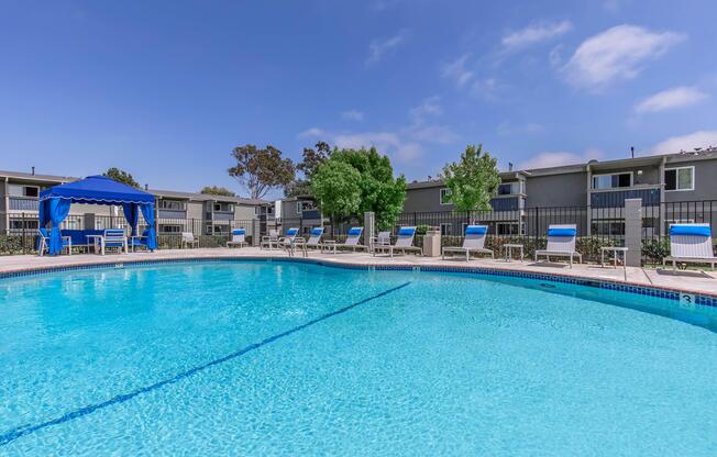 A sunlit swimming pool surrounded by lounge chairs, with a blue canopy on one side. The pool reflects the clear blue sky and is set against a backdrop of landscaped trees and residential buildings. Ideal for relaxation and leisure activities.