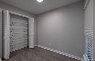 A simple room with light gray walls and wooden flooring. One wall has a window with white blinds, while the opposite wall features a double closet with shelving inside. The room is well-lit by a ceiling light, and the overall space appears empty and ready for decoration.